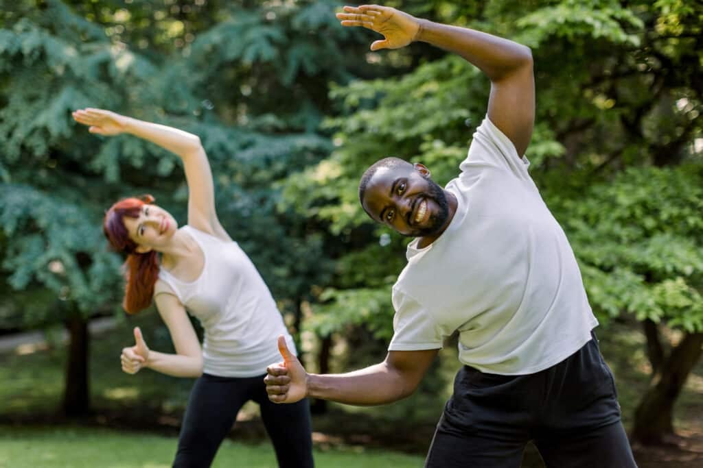 Caucasian woman and African man, fitness couple, training together doing stretching body exercises in the park at sunrise, smiling to camera and showing their thumbs up. Sport strecthing concept.