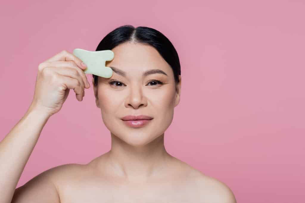 Asian woman with naked shoulders scraping face with jade stone isolated on pink