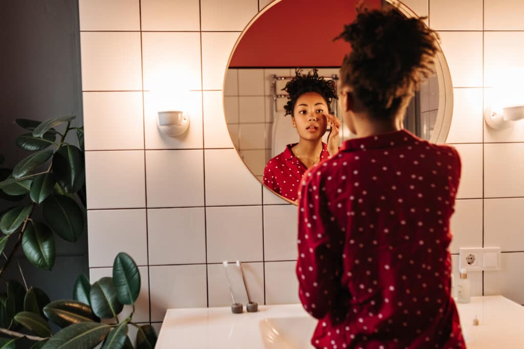 Portrait of curly girl in pajamas, applying cream on her face in front of mirror Portrait of curly girl in pajamas, applying cream on her face in front of mirror
