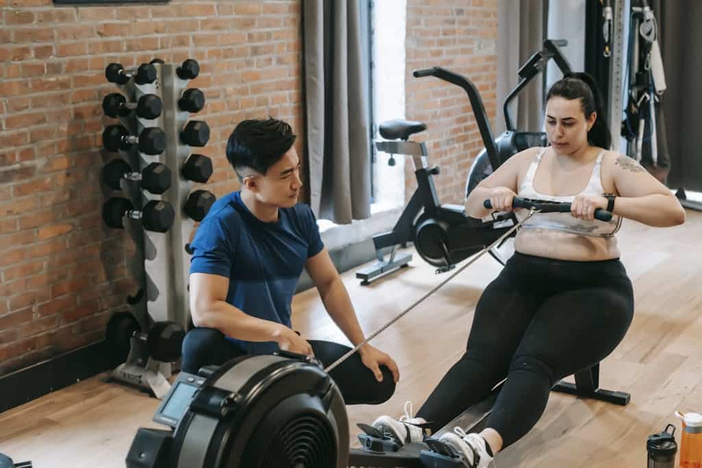 Photo by Andres Ayrton From above of professional ethnic constructor helping young plus size woman working out with rowing machine in sport club