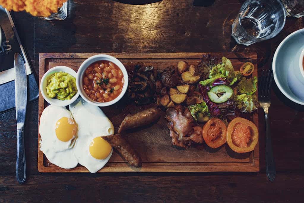 Top view of delicious fried eggs and vegetables with sausages and beans on wooden tray with fork and knife