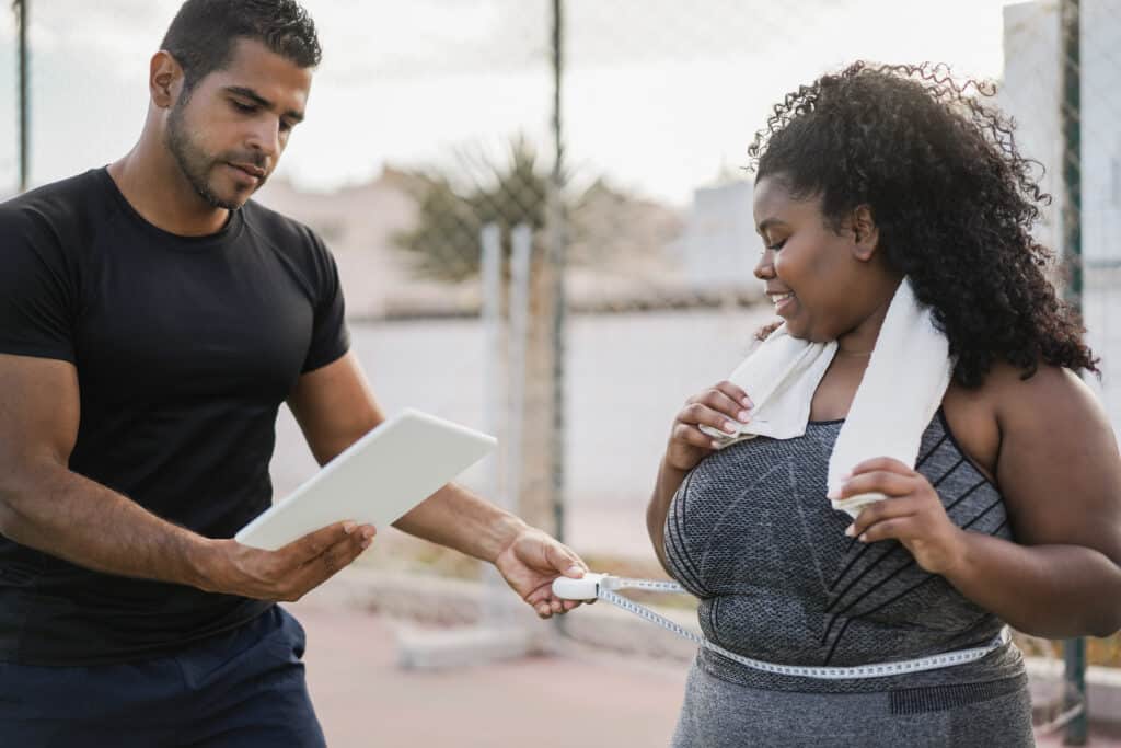 Curvy woman with her personal trainer measuring body waist outdoor - Focus on african girl face Curvy woman with her personal trainer measuring body waist outdoor - Focus on african girl face