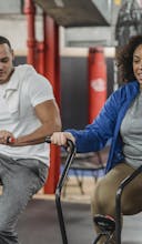 Plus sized African American female in sportswear exercising on stationary bicycle near fit muscular male coach in modern gym