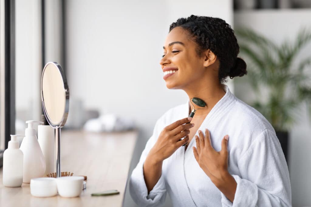 Attractive Young Black Woman Massaging Neck With Greenstone Jade Roller At Home
