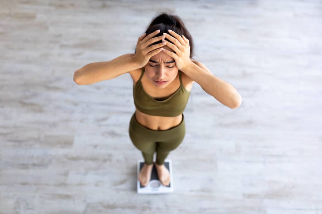 Upset young Indian woman unhappy about her weight, standing on scales, holding head in disappointment, above view