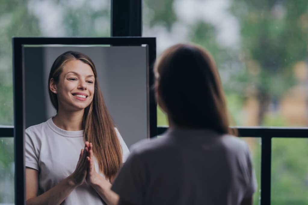 Grateful young brunette hispanic woman in white t-shirt stands at mirror toothy smiles folded hands Grateful young brunette hispanic woman in white t-shirt stands at mirror toothy smiles folded hands