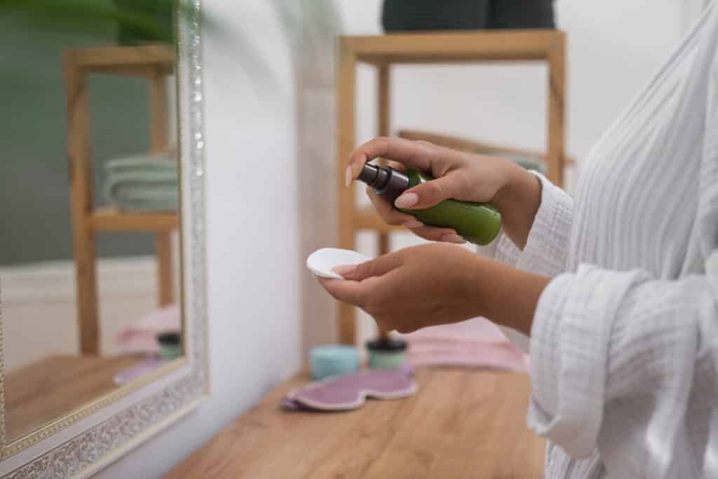 woman in bathroom, skincare regimen, dabs cleansing lotion onto a cotton pad beside the mirror.