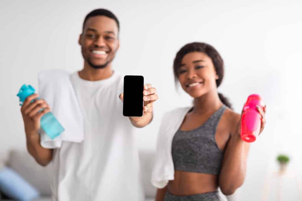 Young athletic black couple holding bottles of water and showing smartphone with empty screen at home, mockup