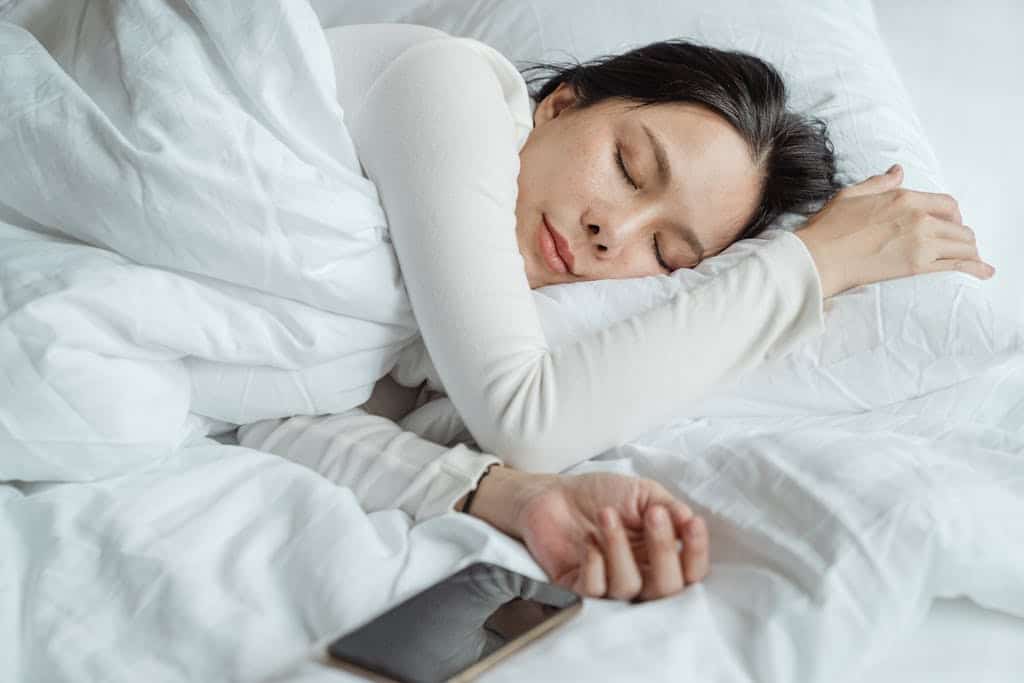 Photo by Ketut Subiyanto Calm Asian female wearing white pajama sleeping in comfortable bed with white sheets near modern mobile phone in morning