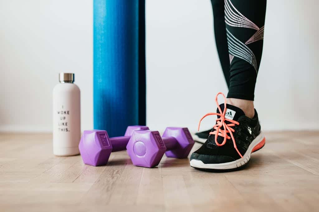 Photo by Karolina Kaboompics Crop sportswoman wearing sneakers and black leggings standing with sport mat on wooden floor near dumbbells and water bottle before exercising indoors