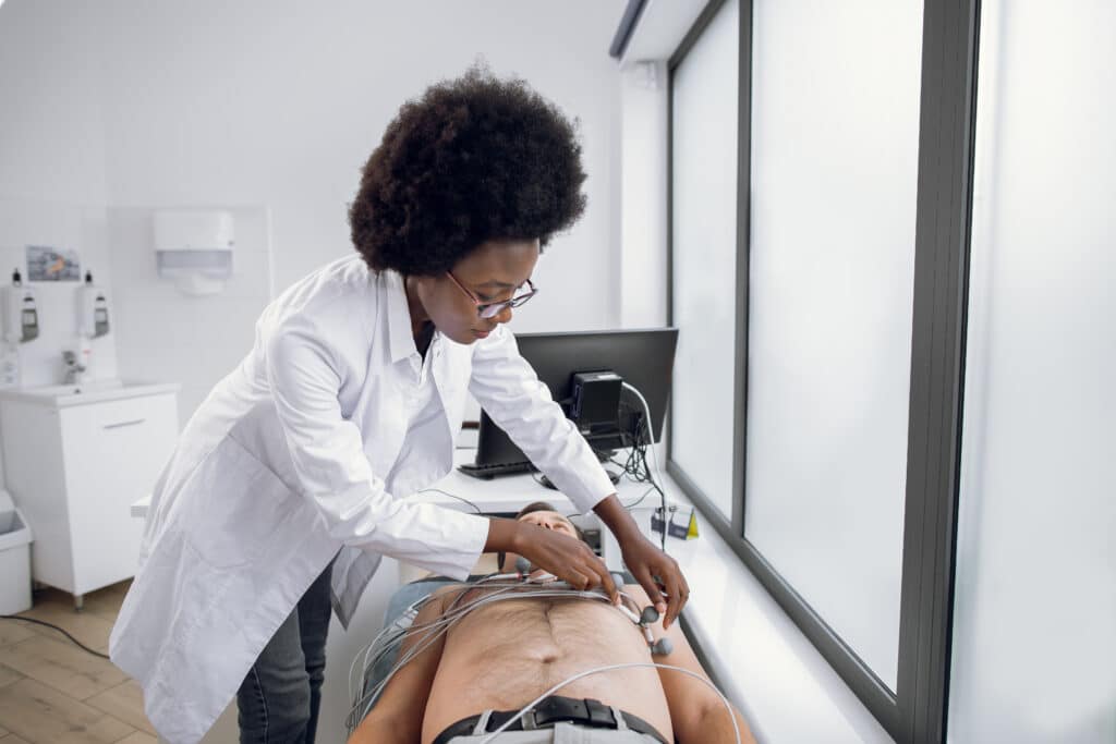 Medicine, health and heart check-up concept. Young afro-american woman doctor with electrocardiogram equipment making cardiogram test to male patient in hospital, fixing electrodes on patient chest.