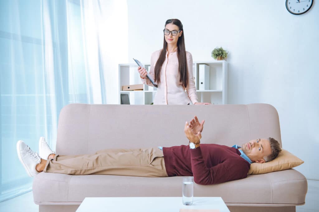 attractive psychotherapist in eyeglasses holding clipboard and smiling at camera while patient lying