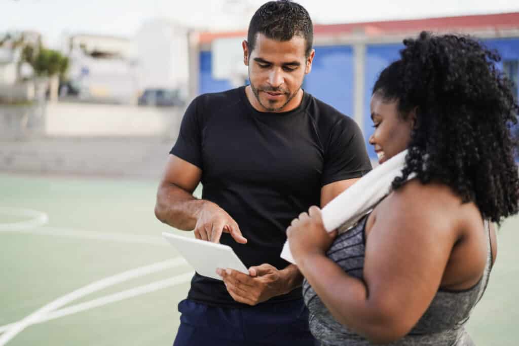 Curvy woman talking with her personal trainer while checking tech clipboard outdoor - Focus on trainer
