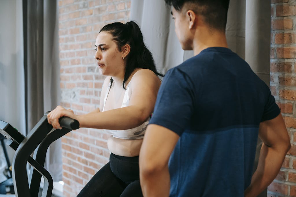 Photo by Andres Ayrton Side view of serious concentrated young obese female doing weight lose exercise on cycling equipment during cardio training with crop ethnic male personal coach