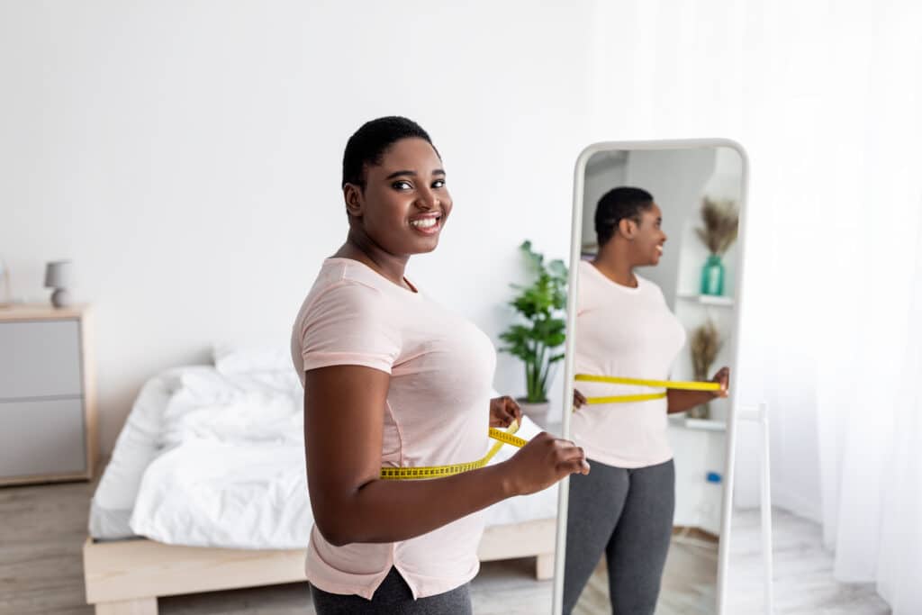 Curvy Afro woman measuring waist with tape near mirror, satisfied with weight loss result, achieving slimming success Curvy Afro woman measuring waist with tape near mirror, satisfied with weight loss result, achieving slimming success