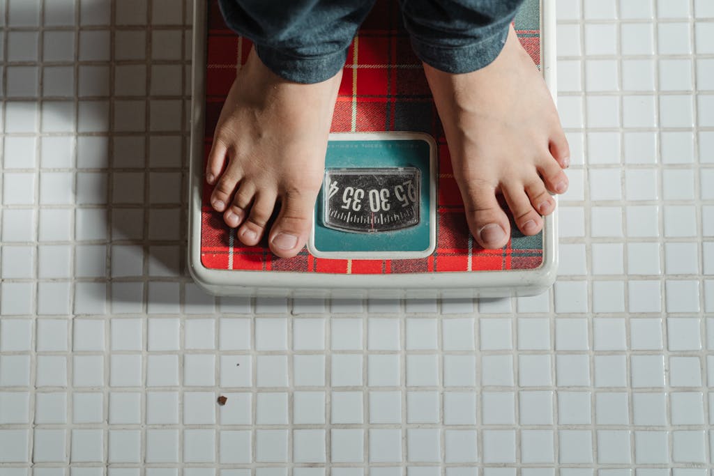 Photo by Ketut Subiyanto From above crop anonymous barefoot child in jeans standing on weigh scales on tiled floor of bathroom