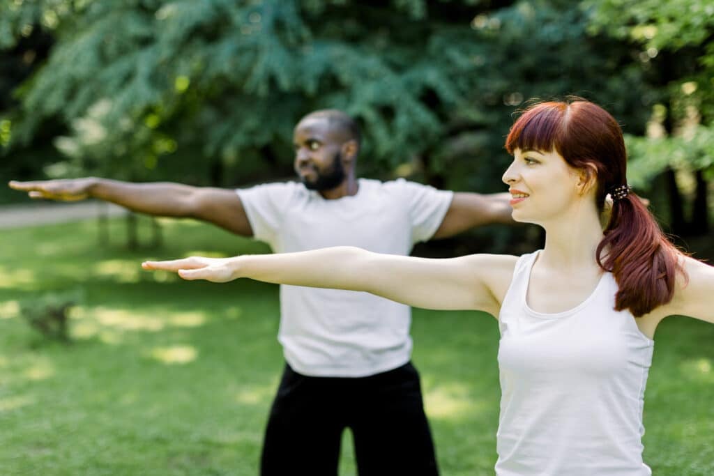 Concept of healthy family lifestyle and yoga at park. Young multiethnic couple exercising outdoors with arms outstretched, looking ahead. Focus on pretty Caucasian girl.
