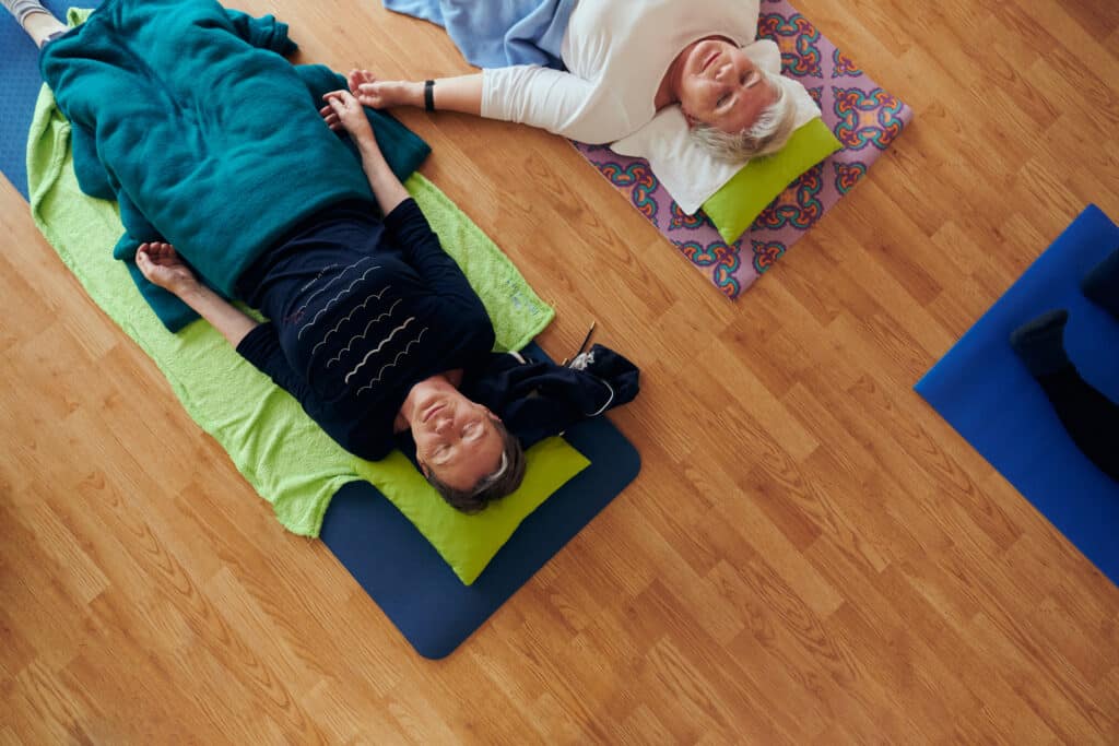 Top view of group of senior women engage in various yoga exercises, including neck, back, and leg stretches, under the guidance of a trainer in a sunlit space, promoting well-being and harmony