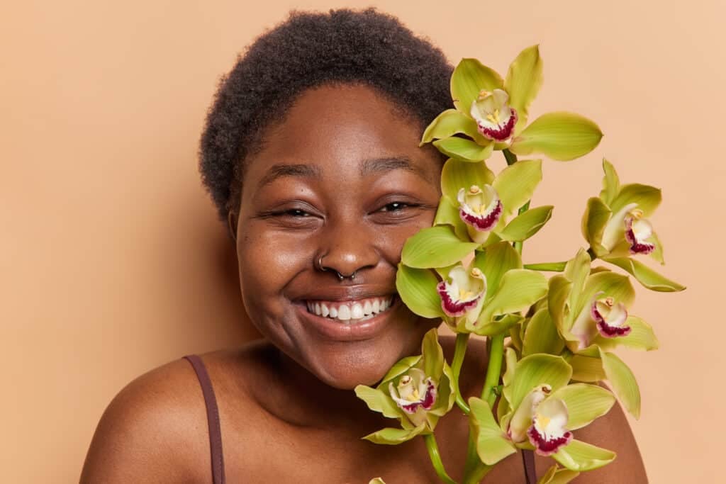 Horizontal shot of cheerful Afro woman has short hair and dark skin smiles toothily holds flower near face going to undergo beauty procedures stands bare shoulders isolated over brown background