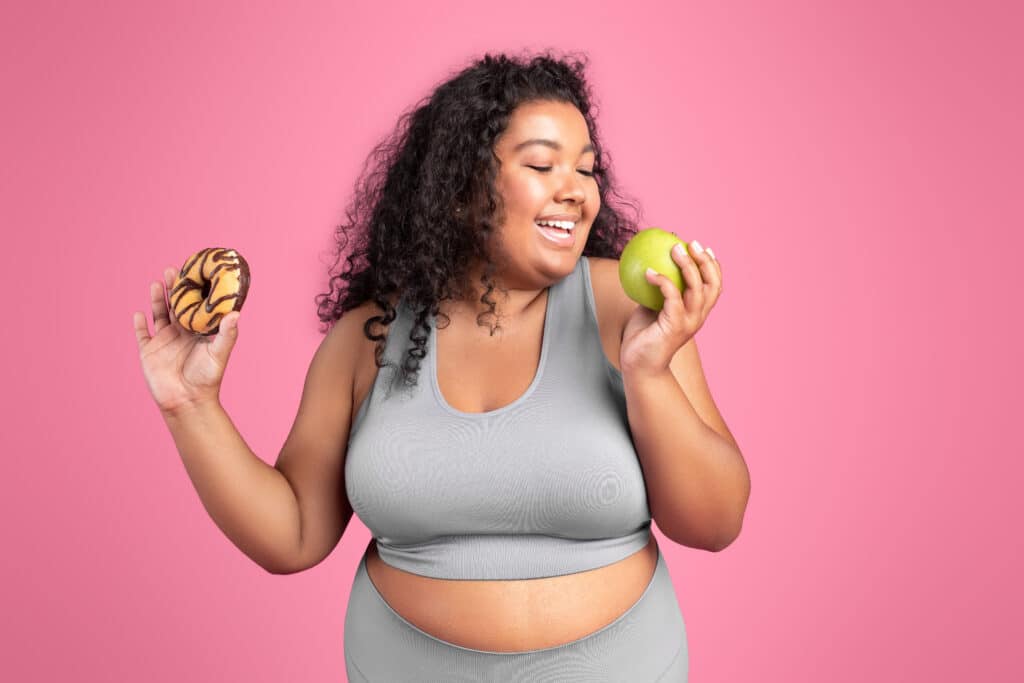 Young black body positive lady in sportswear holding donut and biting apple, choosing healthy food, pink background Young black body positive lady in sportswear holding donut and biting apple, choosing healthy food, pink background